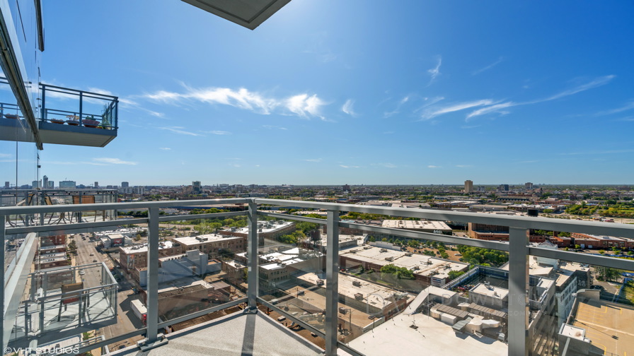View from a balcony with a glass railing overlooking an urban cityscape. Buildings and roads are visible under a clear blue sky.
