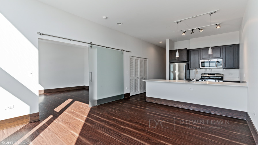 Modern kitchen with dark cabinets, stainless steel appliances, sliding door leading to another room, and an open area with dark wood flooring and white walls.