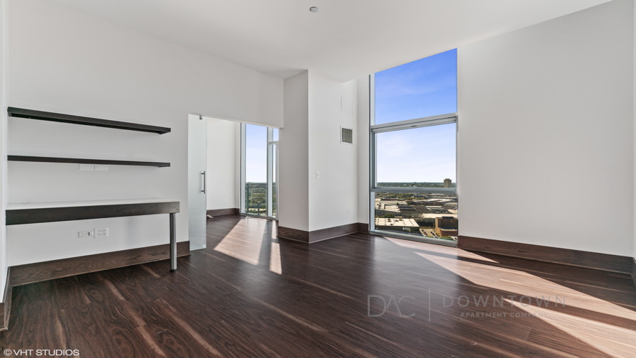 Sunlit room with dark wood flooring, white walls, and large windows overlooking a cityscape. Built-in dark shelves are seen on the left, and an open doorway leads to another room.
