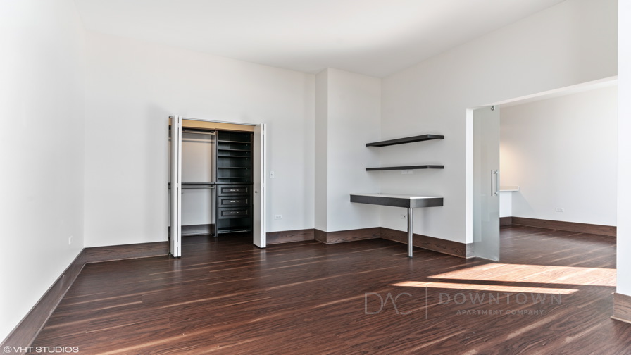 Empty room with hardwood floor, open closet with shelving, wall-mounted shelves on the right, large doorway leading to another room, and white walls.