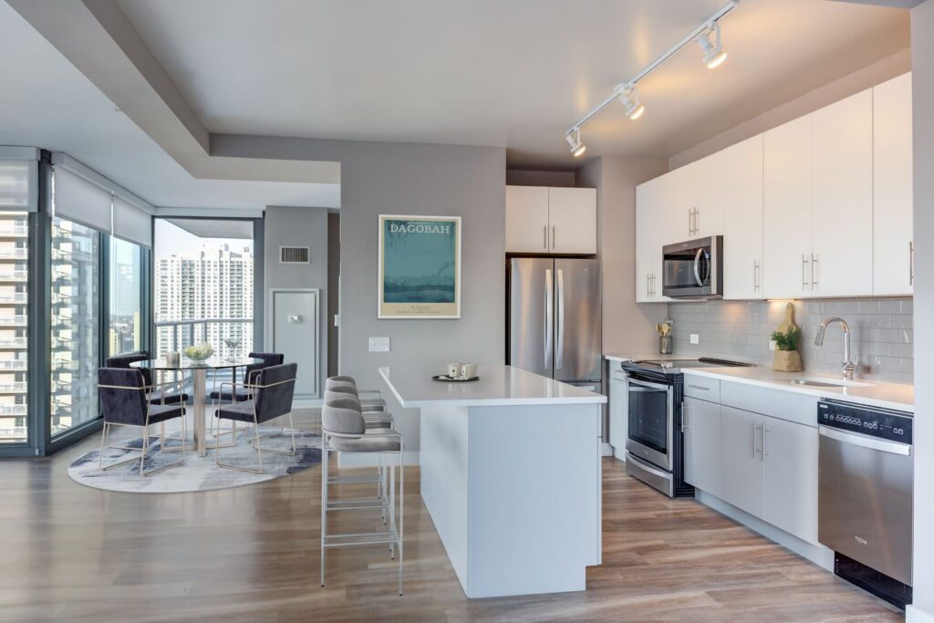 Modern apartment kitchen with white cabinets, stainless steel appliances, an island with barstools, and a dining area near large windows with a city view.