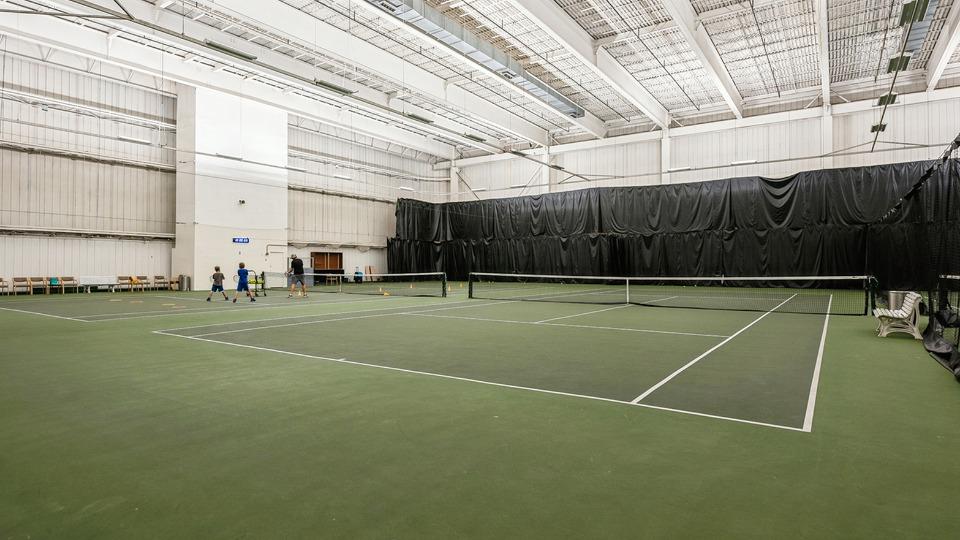 Indoor tennis court with green flooring, net in middle, and large black curtains. Two people stand near a white wall on the left side.