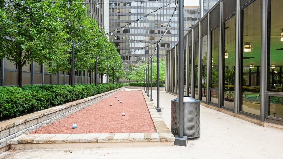 Outdoor bocce ball court with red gravel, surrounded by greenery and string lights, next to a building with glass windows.