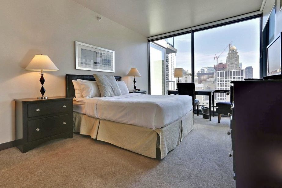 Modern hotel room with a large bed, nightstands with lamps, and a chair by the window offering a cityscape view with skyscrapers and a crane in the background.