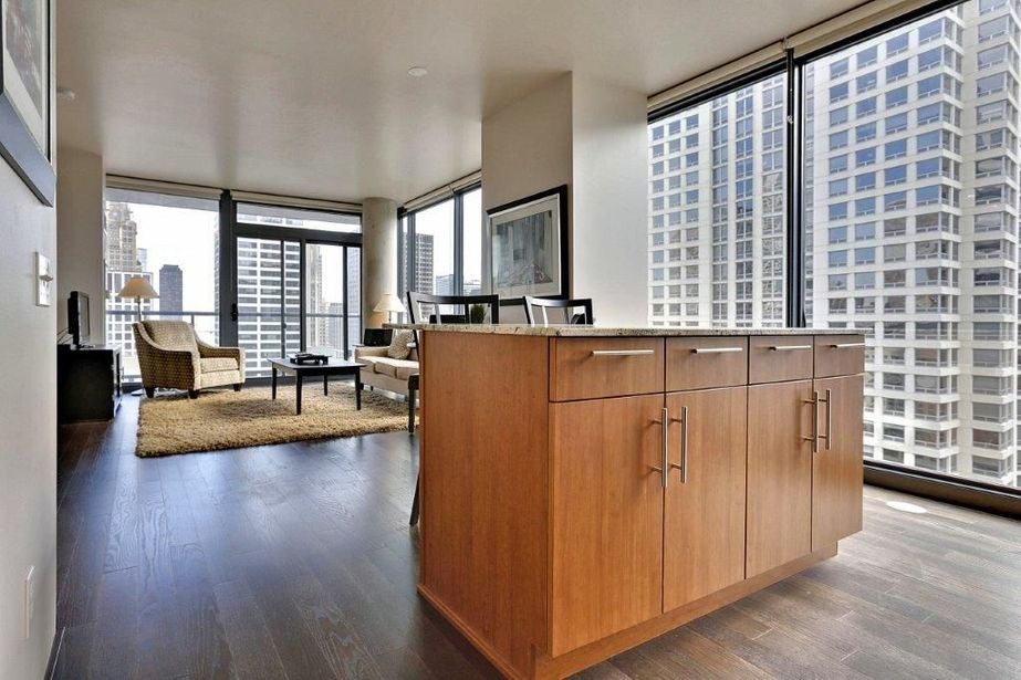 Modern apartment interior with wooden kitchen island, large windows, and city view. Living area has a rug, chairs, and a table.