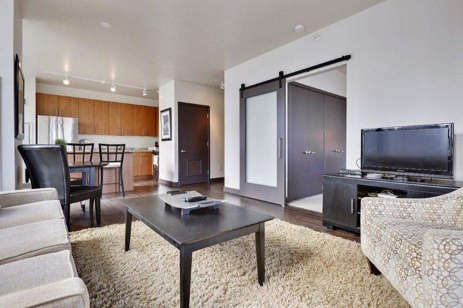 Modern living room with a beige sofa, patterned armchair, black coffee table, TV, and a rug. Kitchen with wood cabinets and bar stools in the background. Sliding barn door visible.