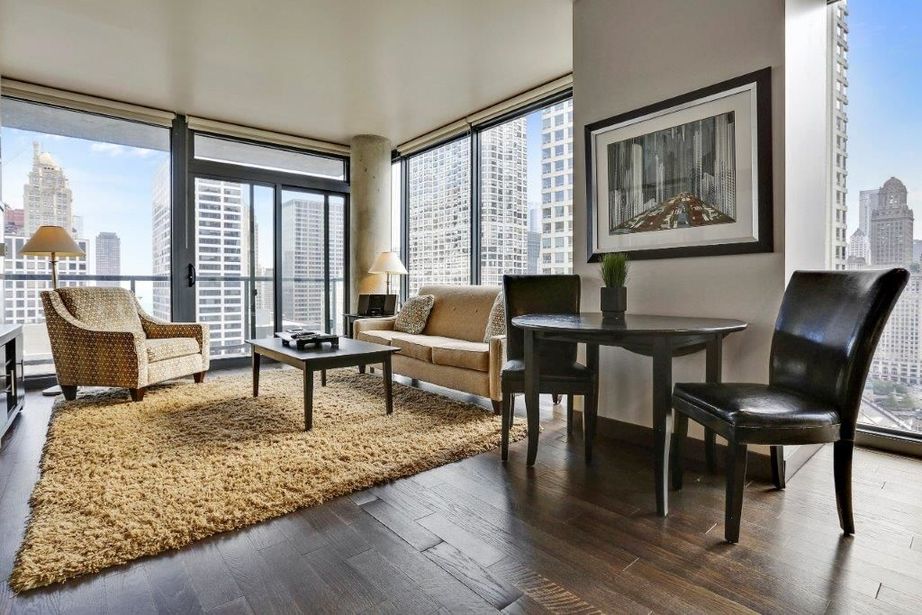 A modern living room with large windows, two chairs, a sofa, a coffee table, and a round dining table with two chairs. A shaggy rug covers the wooden floor, and skyscrapers are visible outside.
