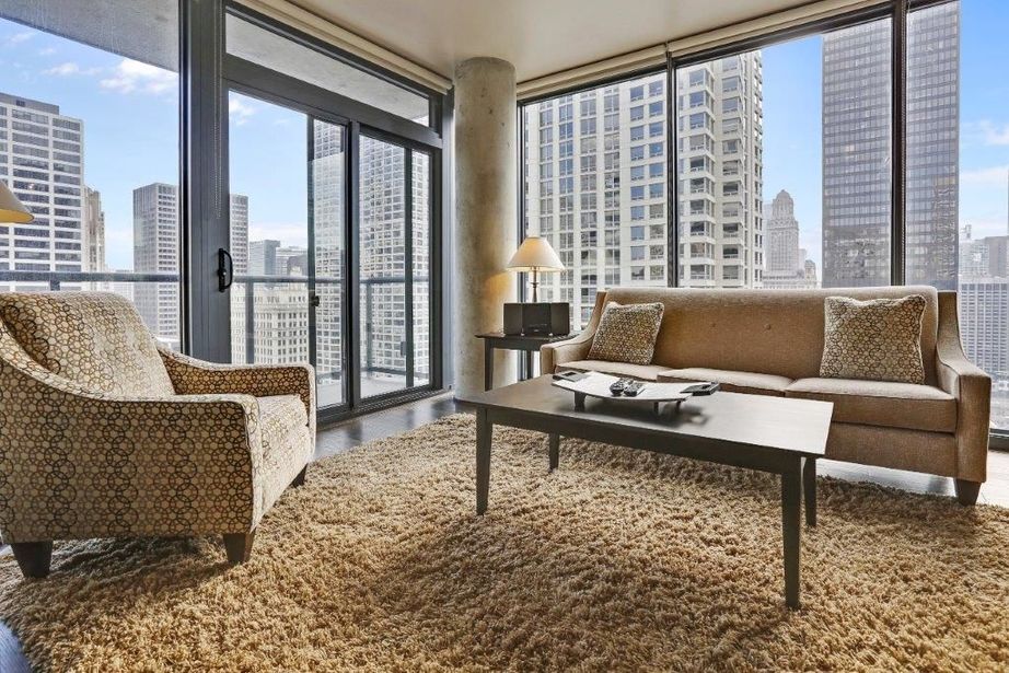 Modern living room with large windows showcasing cityscape. Features a beige sofa, patterned armchair, coffee table, and textured rug.