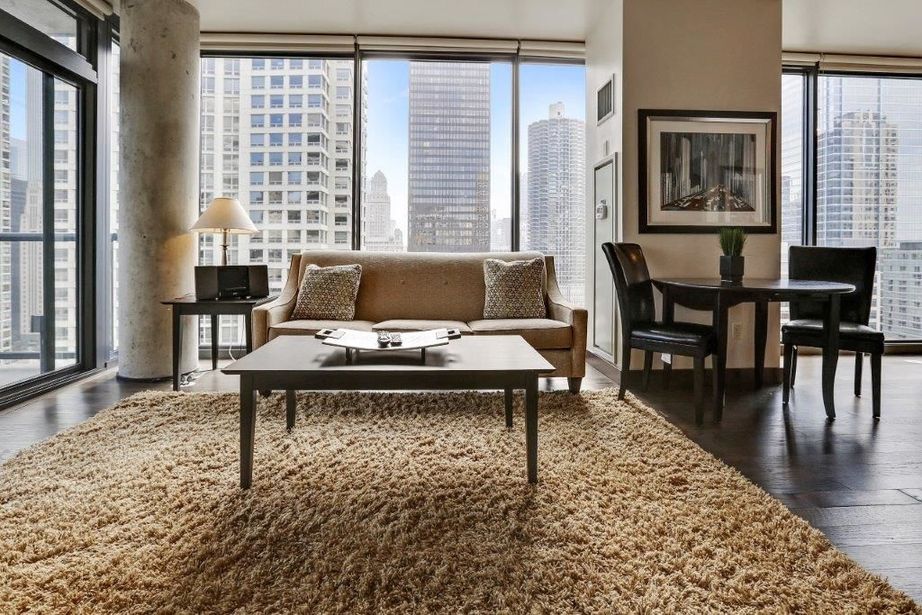 Modern living room with large windows overlooking a cityscape. Features a beige sofa, a wooden coffee table, a rug, and a dining set. Natural light fills the space.