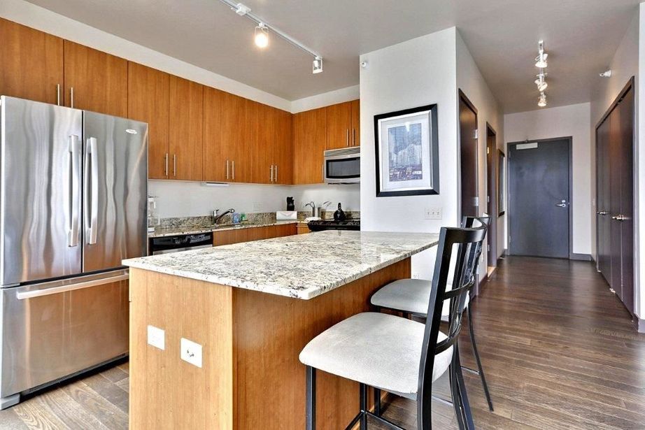 Modern kitchen with wooden cabinets, granite countertops, stainless steel appliances, and a light fixture. An island with two high chairs is in the center, and framed artwork is on the wall.