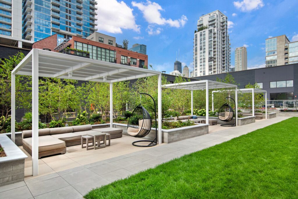 Rooftop lounge area with modern pergolas, seating, and hanging chairs, surrounded by greenery and city buildings in the background under a blue sky.