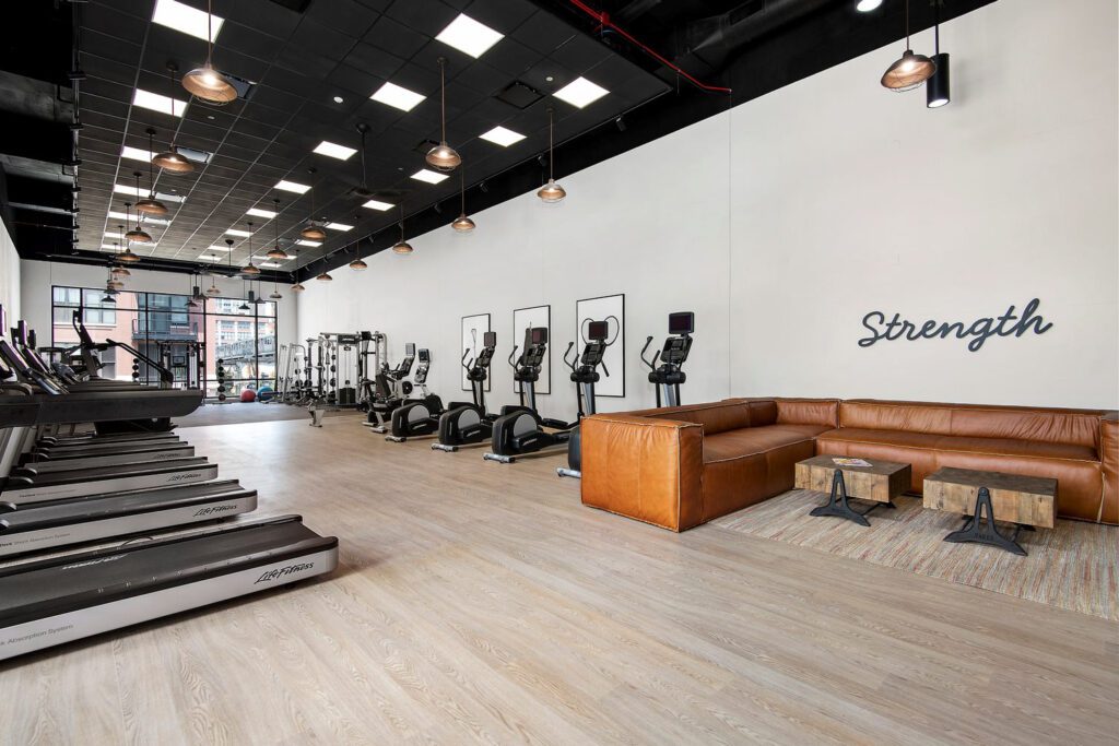 Modern gym interior with treadmills, exercise bikes, and a row of ellipticals on one side. A brown leather seating area with a rug and small tables is on the right. The word "Strength" is on the wall.
