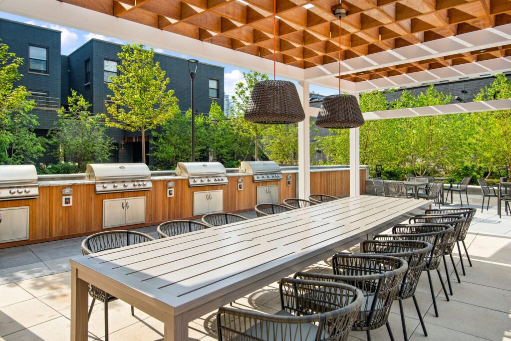 Outdoor dining area with a long table, wicker chairs, a wooden pergola, hanging lights, and four adjacent stainless steel grills. Trees and modern buildings are in the background.