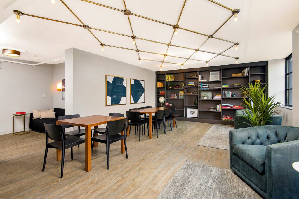Modern lounge area with wooden tables, black chairs, teal sofa, and built-in bookshelves. Geometric artworks adorn the wall, and a potted plant adds greenery. Ceiling lighting features gold fixtures.