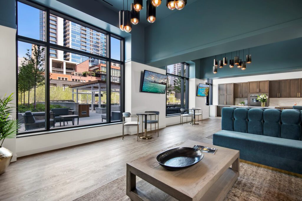 Modern lounge area with large windows, blue ceiling, and contemporary decor. Includes seating, hanging lights, and wall-mounted TVs. Outdoor view of buildings and greenery visible.