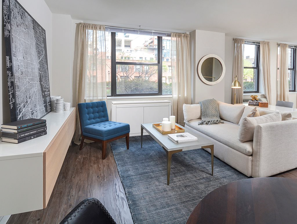 Modern living room with a beige sofa, blue armchair, white coffee table, and wall-mounted TV. Large windows with light curtains and a city view. Decor includes books, candles, and a round mirror.