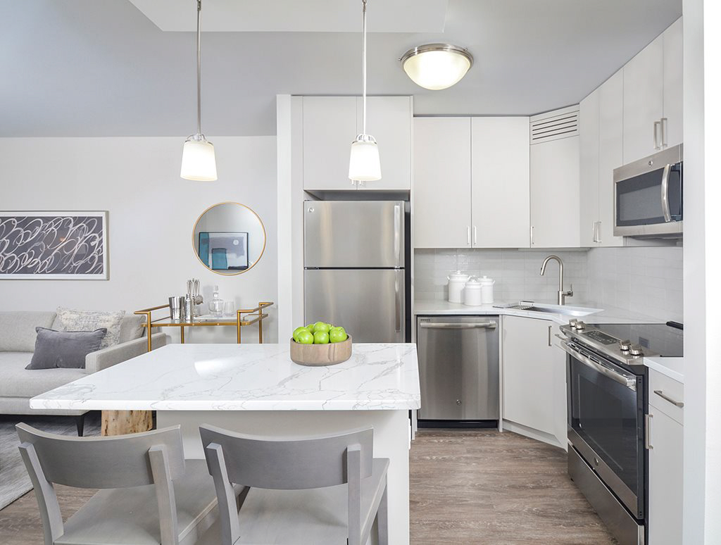 Modern kitchen with white cabinets, stainless steel appliances, and a marble island. Two gray chairs are at the island, and there is a bowl of green apples. The living area is visible in the background.