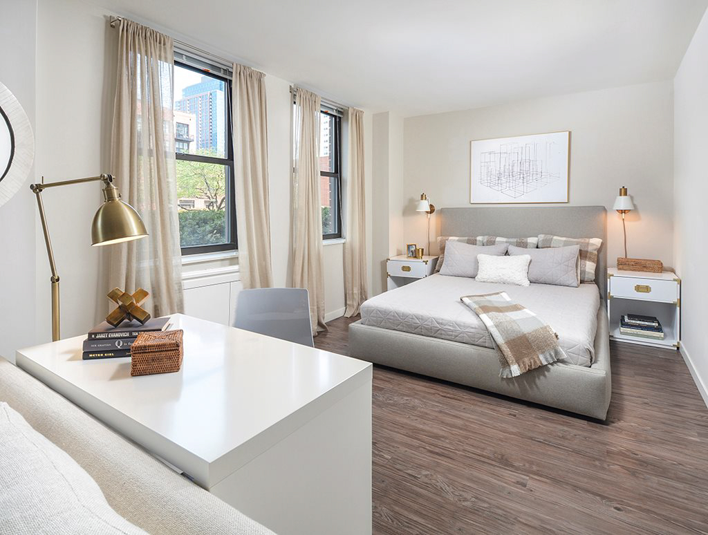 A modern bedroom with a bed, two bedside tables with lamps, a desk with a chair, and large windows with beige curtains.