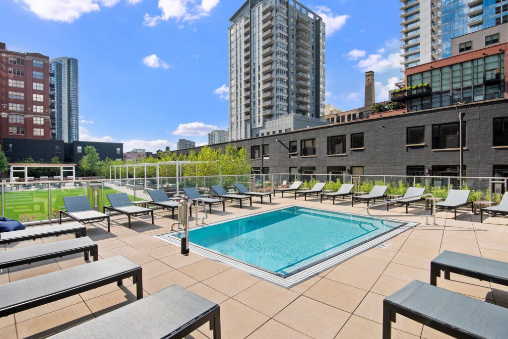 Rooftop pool area with lounge chairs on a sunny day. Surrounded by modern high-rise buildings and a clear blue sky.