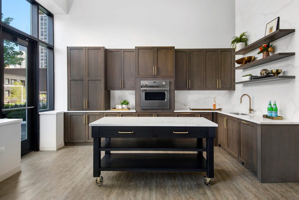 Modern kitchen with dark wooden cabinets, a marble-topped island on wheels, stainless steel oven, open shelves with decor, and a view of large windows showing an outdoor scene.