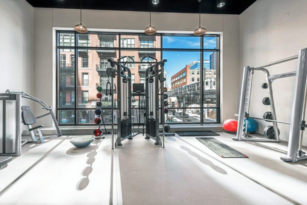 A modern gym with various exercise equipment, large windows showing an urban view, and light streaming in.