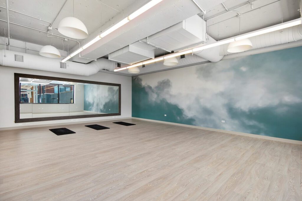 Empty fitness studio with wooden floors, large mirrors along one wall, and a mural of clouds on the opposite wall. Ceiling features exposed ducts and hanging lights. Three black mats on the floor.