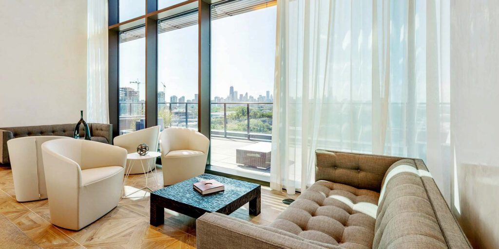 Modern lounge with beige armchairs, a tufted sofa, and glass wall overlooking a cityscape. Decor includes a black vase, spherical sculpture, and a book on a square coffee table.