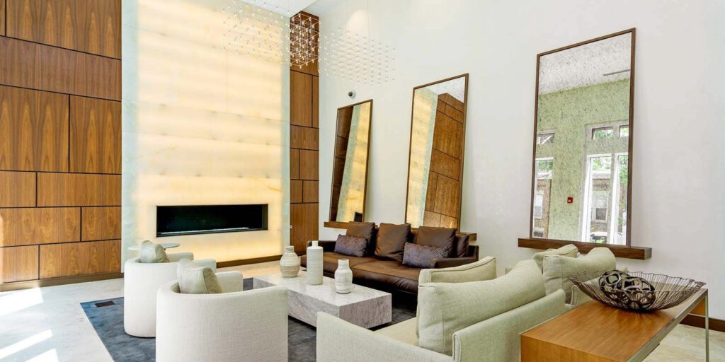 Modern living room with high ceiling, featuring a large fireplace, wooden paneling, beige sofas, white armchairs, and tall mirrors. Decor includes ceramic vases and a wire bowl on tables.