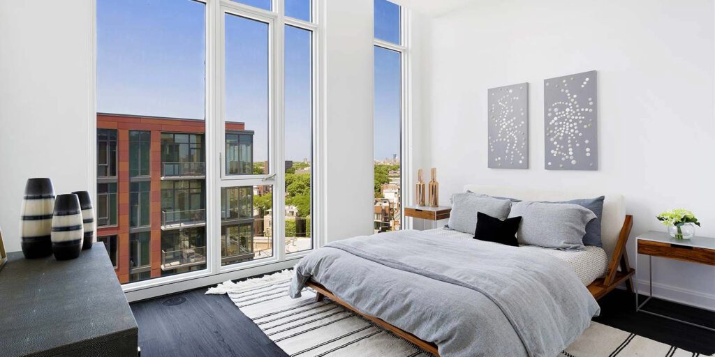 Modern bedroom with large windows, gray bedding, and two abstract artworks above the bed. A wooden nightstand with decor is on the right, and a striped rug covers the dark floor.