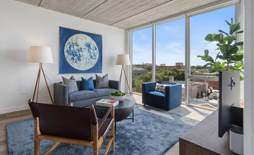 Modern living room with large windows, blue and gray decor, a sofa, armchair, two lamps, and wall art of a celestial design. A potted plant and books are also visible.