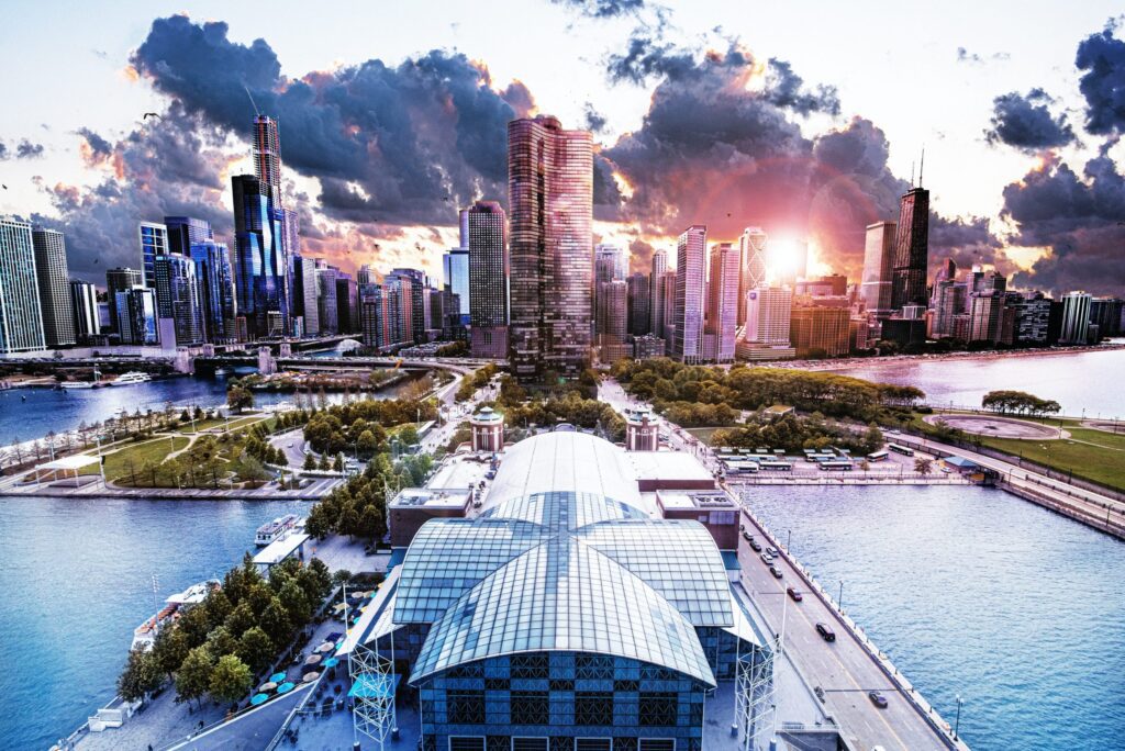 Aerial view of a city skyline with tall buildings at sunset, featuring a waterfront area and a large glass structure in the foreground.