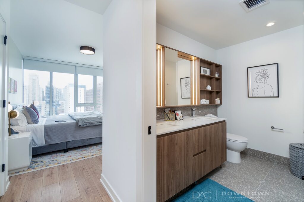 Modern apartment interior with a view of the city skyline. Bedroom features a bed and large windows. Bathroom includes a double sink, toilet, and wall art.