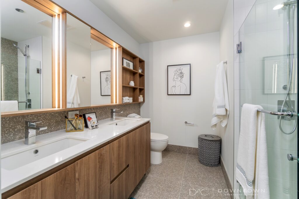 Modern bathroom with a double sink vanity, wood accents, a shower with glass doors, and wall art above the toilet. Towels are neatly hung, and open shelves hold decorative items.