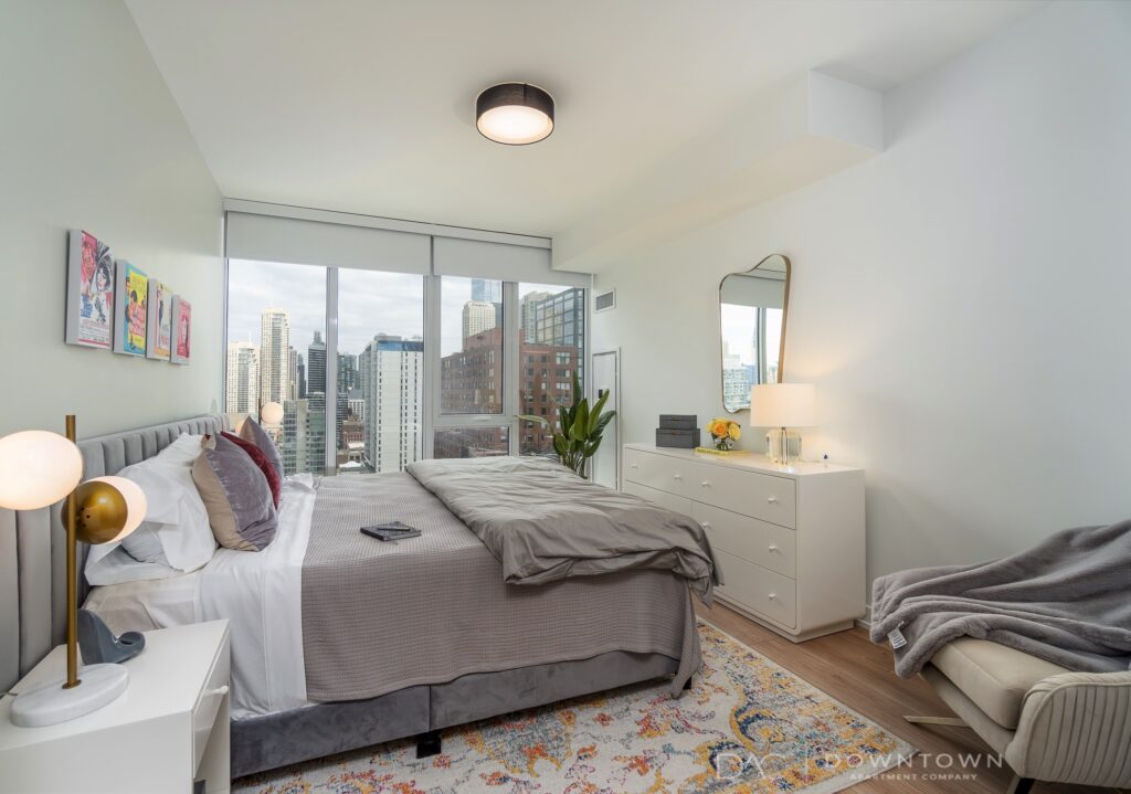 Bright bedroom with a large window view of city buildings. Features a bed with gray bedding, a dresser with a mirror, a side table with a lamp, and wall art.