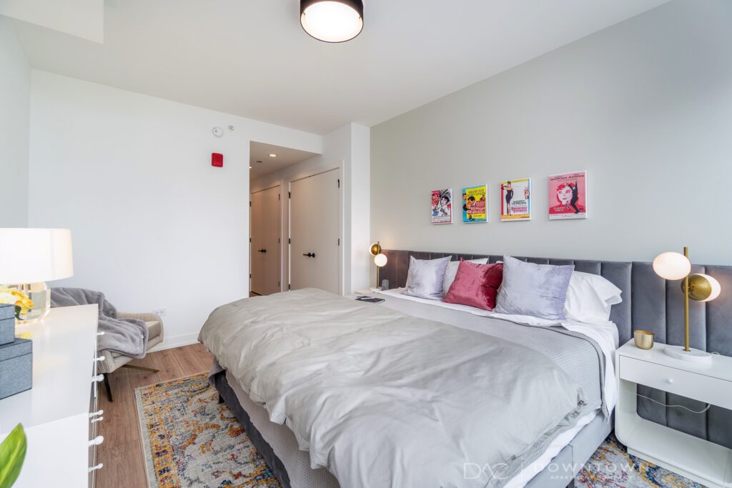 A modern bedroom with a large bed, white linens, and colorful pillows. Artwork hangs above the bed. A dresser and a lamp are on the left. Light gray walls and wood flooring complete the space.
