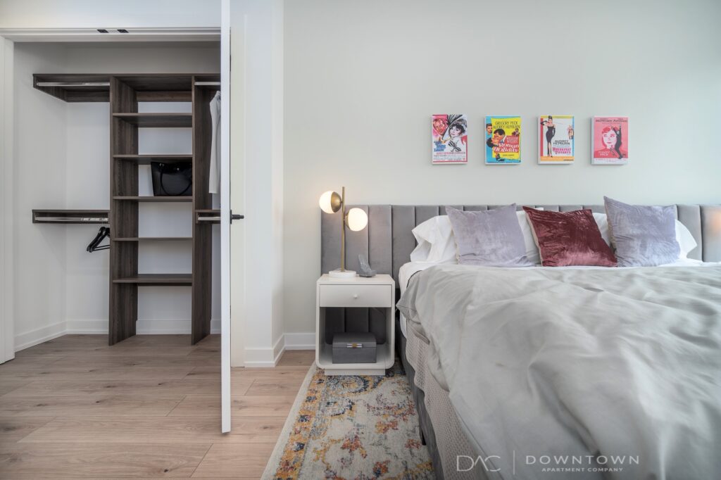Modern bedroom with an open closet. The room features a neatly made bed, bedside table with lamp, and colorful wall art above. Floor is light wood. Logo on the wall reads "Downtown Apartment Company.