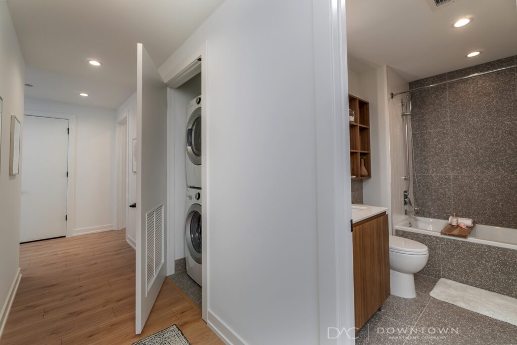 Modern bathroom with a bathtub, showerhead, and vanity sink. Laundry area with stacked washer and dryer is visible in an open closet to the left. Wood flooring and white walls.
