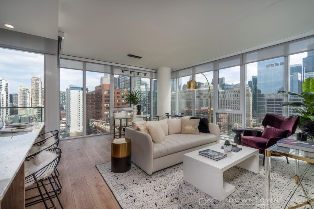 Modern city apartment with floor-to-ceiling windows, white sofa, purple chair, and contemporary decor. Overlooks urban buildings and skyscrapers. Natural light fills the space.