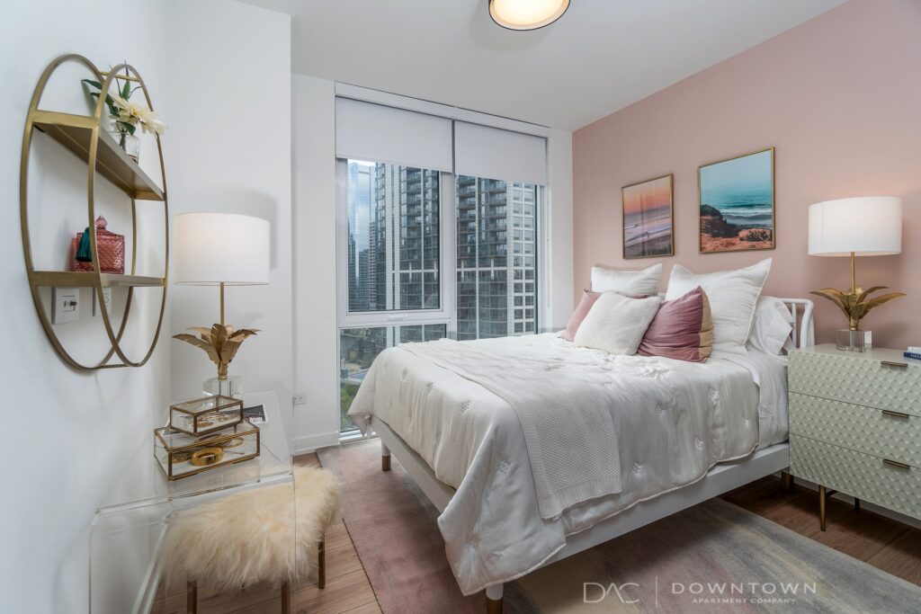 A modern bedroom with a plush bed, two side tables, and decorative items. Large window with city view, wall art, and a round shelf. Room has soft lighting and contemporary decor.