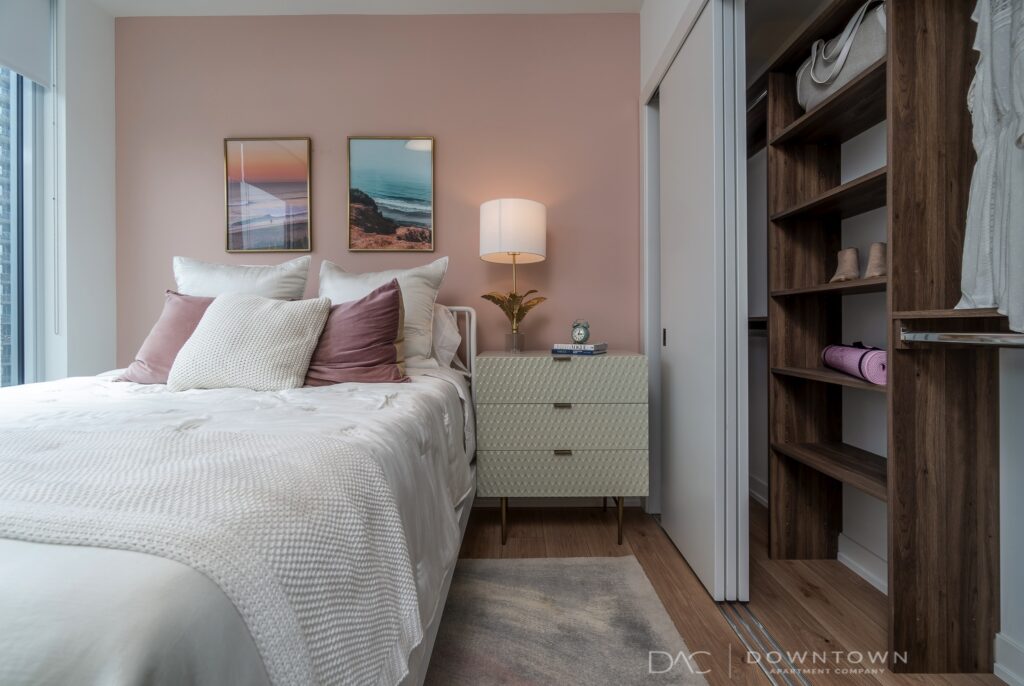 Modern bedroom with a neatly made bed, pink accent wall, two framed pictures, a bedside table with a lamp, and an open closet with shelves displaying clothing and shoes.