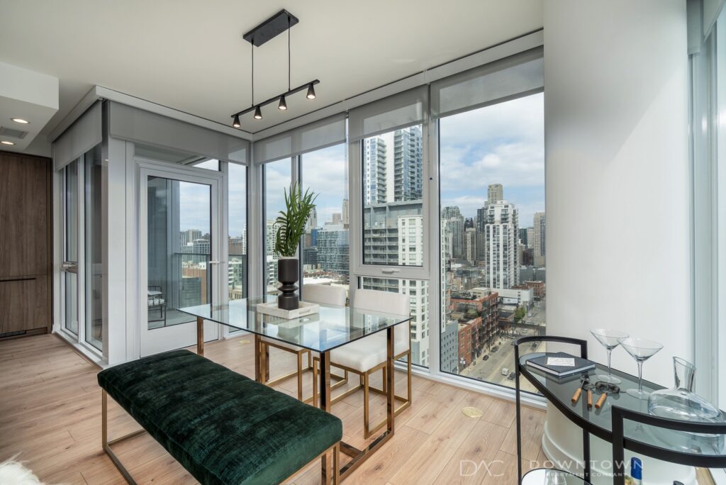 Modern dining area with glass table, green bench, and cityscape view through large windows. Martini glasses and a tray are on the table.