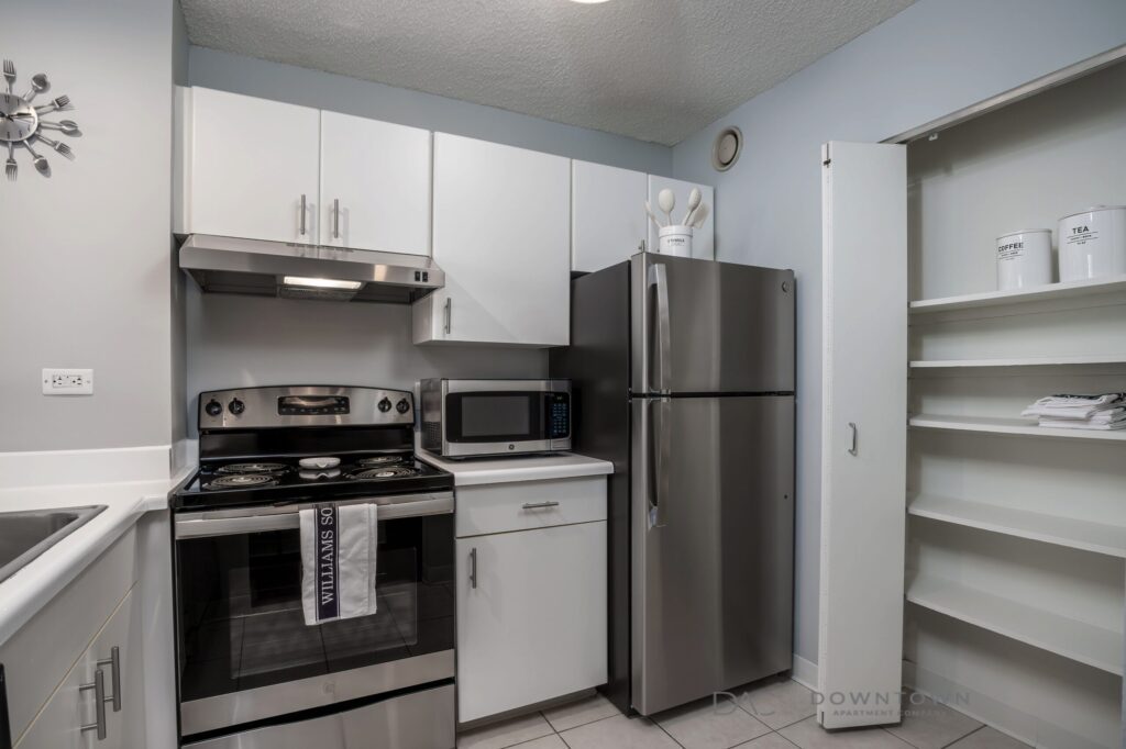 Modern kitchen with stainless steel appliances, white cabinets, a microwave, and an electric stove. Open shelves on the right hold cups, tins, and towels.