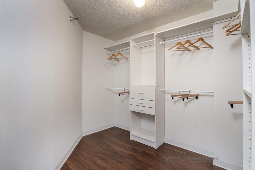 Empty walk-in closet with white shelves, hanging rods, and a light fixture. Brown wooden floor and several wooden hangers are visible.