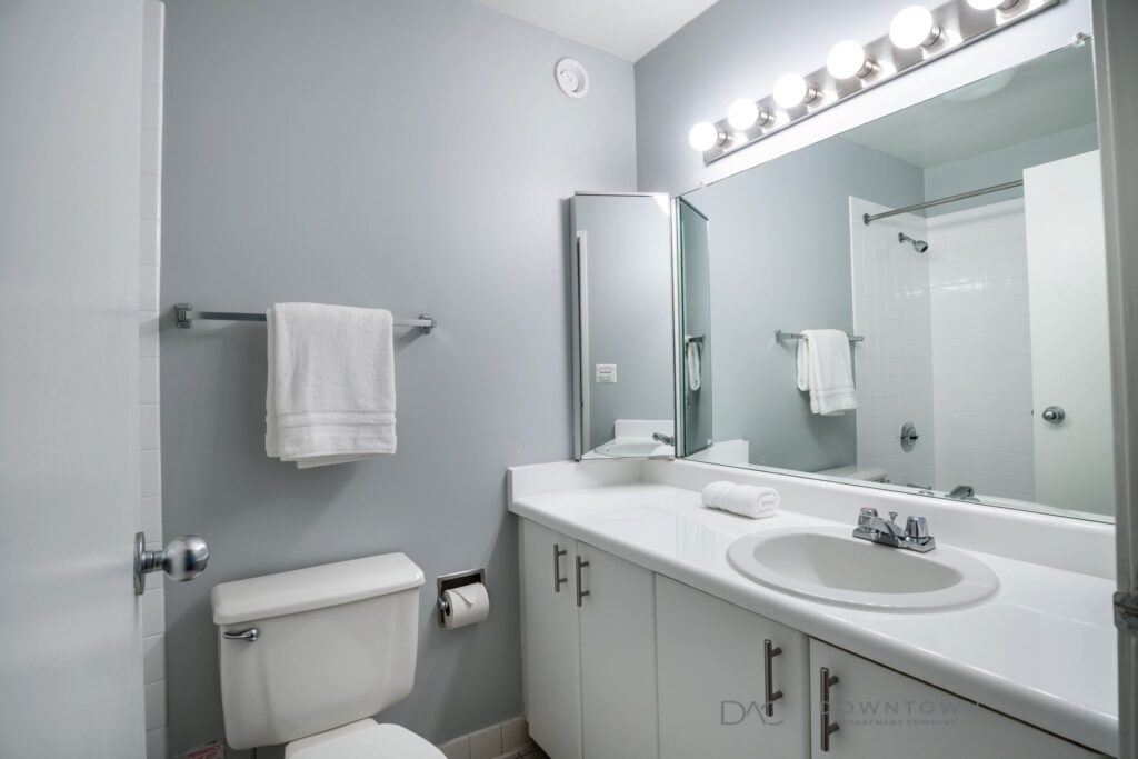 Modern bathroom with light gray walls, white countertop, and large mirror. Features a sink, toilet, towel rack, and a shower with a curtain. Bright vanity lights above the mirror.
