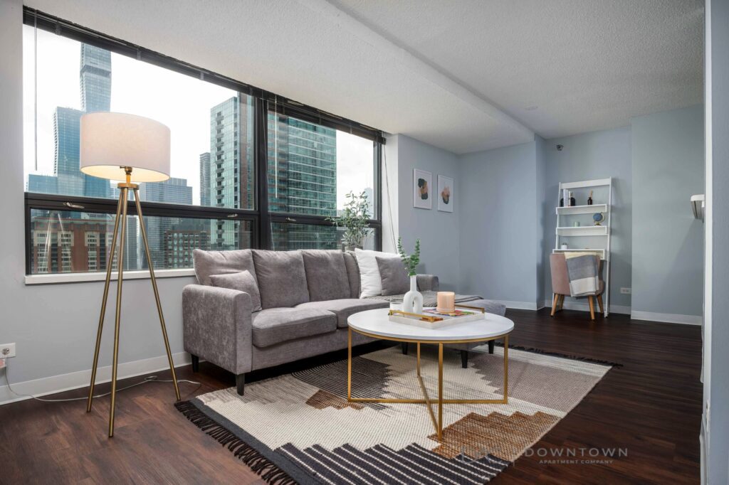 Modern living room with a gray couch, round coffee table, and floor lamp on a geometric rug. Large windows reveal a cityscape. A bookshelf and small desk are in the corner.