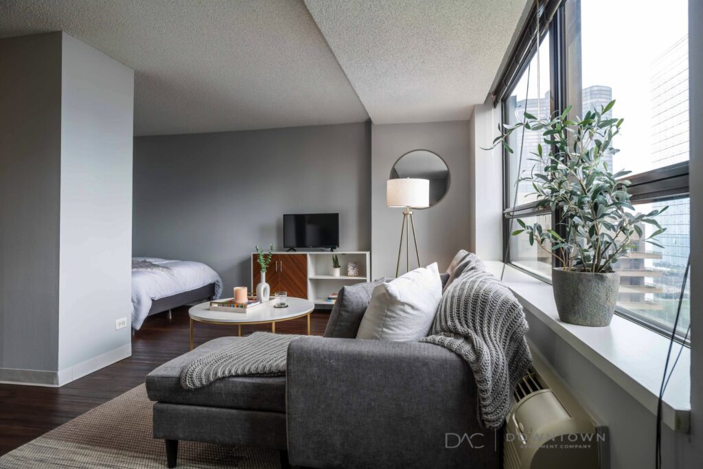 Modern apartment living room with a gray sectional sofa, coffee table, TV on a stand, and a floor lamp. Bedroom partially visible through doorway. Large window offers a city view.