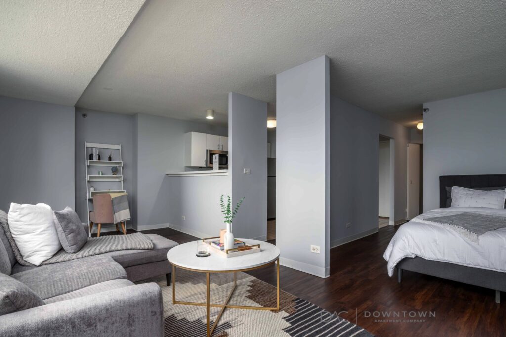 Modern studio apartment with an open layout. Features a gray sofa, round coffee table, and a bed. Kitchen area with bar seating is visible. Walls and decor are in neutral tones.