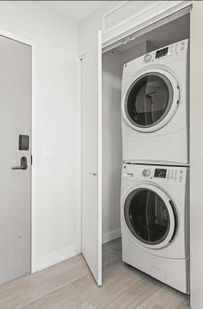 Stacked washer and dryer in a white closet with an open door, next to a closed gray entry door on a light wood floor.