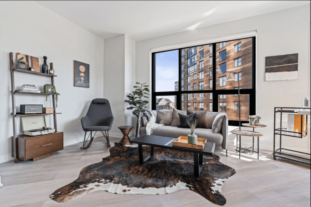 Modern living room with a large window view of an apartment building. Features a gray sofa, black chair, wooden furniture, potted plants, and a cowhide rug on light wooden flooring.