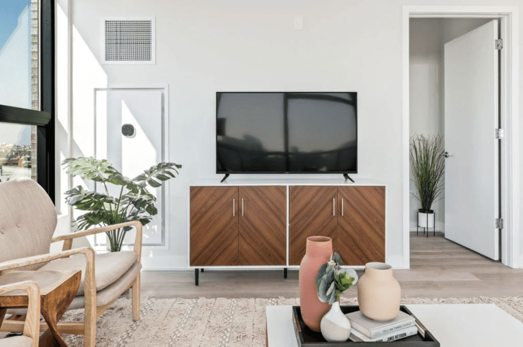 Living room with a flat-screen TV on a wooden stand, a beige chair, potted plants, and decorative vases on a coffee table. Door slightly open in the background. Natural light from window.